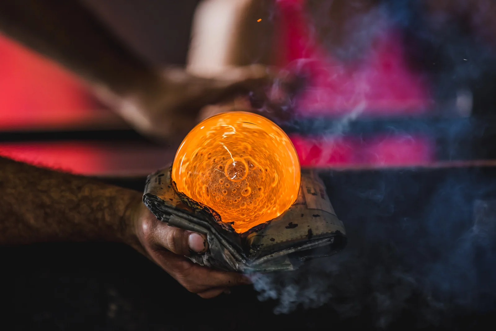 Hands shape glowing orange glass with folded material as smoke rises around it.