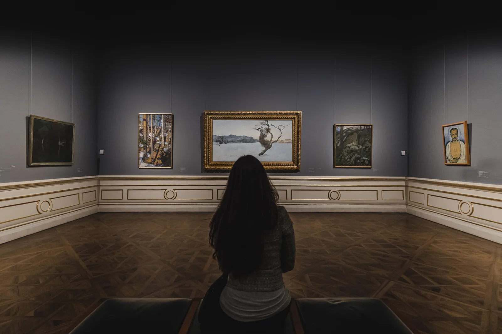 A woman sits with her back to the camera in a symmetrical art gallery, facing a wall of paintings.