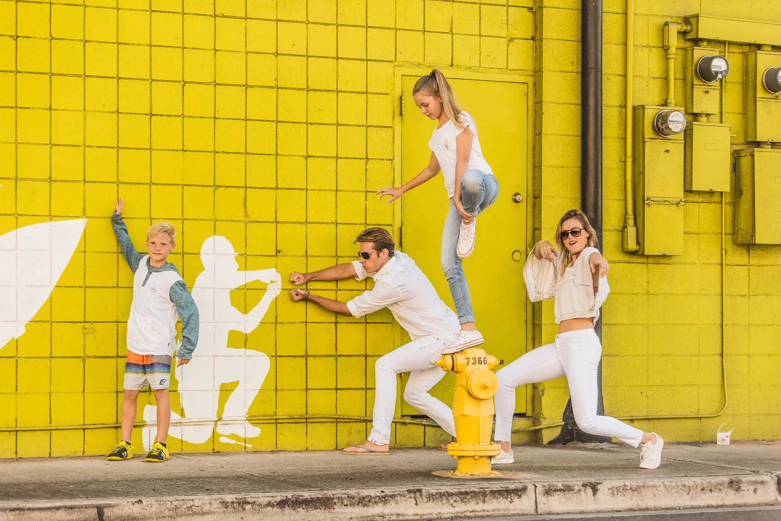 Four people strike playful poses beside a painted silhouette on a bright yellow tiled wall.