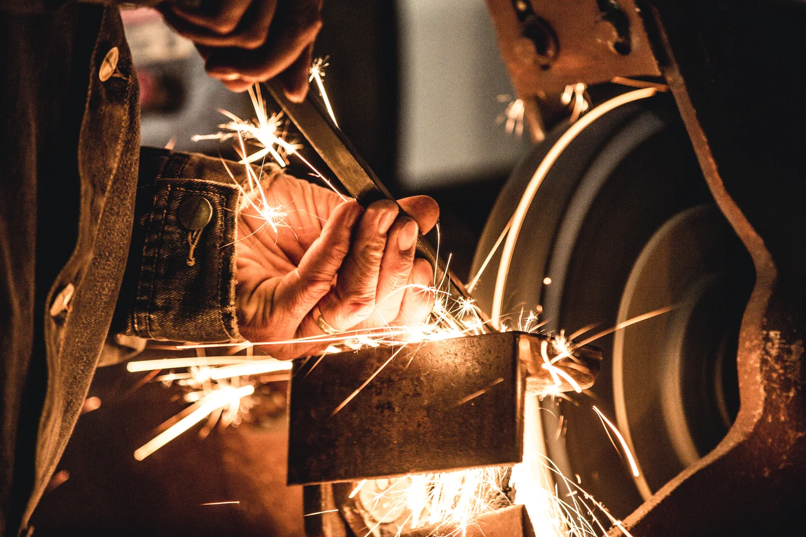 An artisan sharpens a tool against a grinding wheel, sending a fan of orange sparks across the workshop.