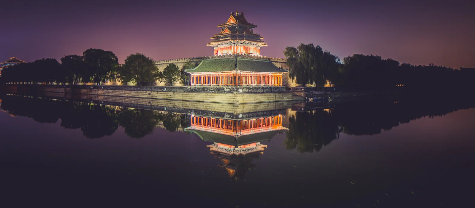 An illuminated traditional pavilion is reflected almost perfectly in dark water at night.