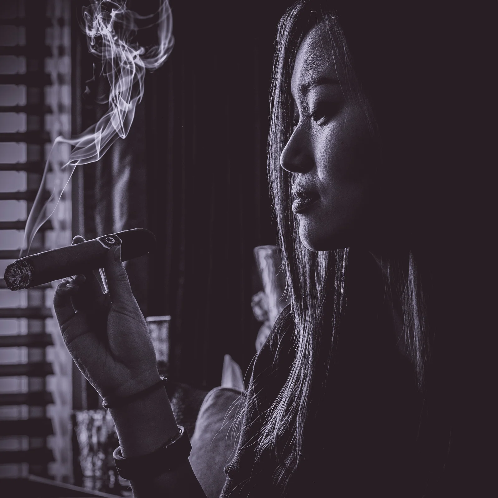 In black and white, a woman holds a cigar beside a window as smoke curls through the light.