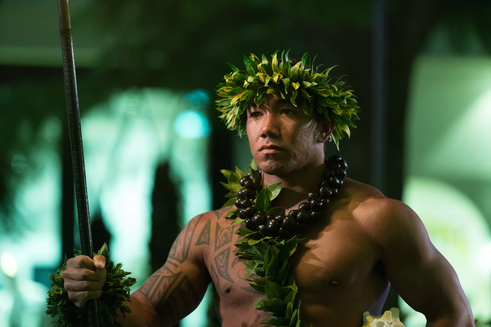 A bare-chested performer wearing a leafy crown and necklace holds a staff against green light.